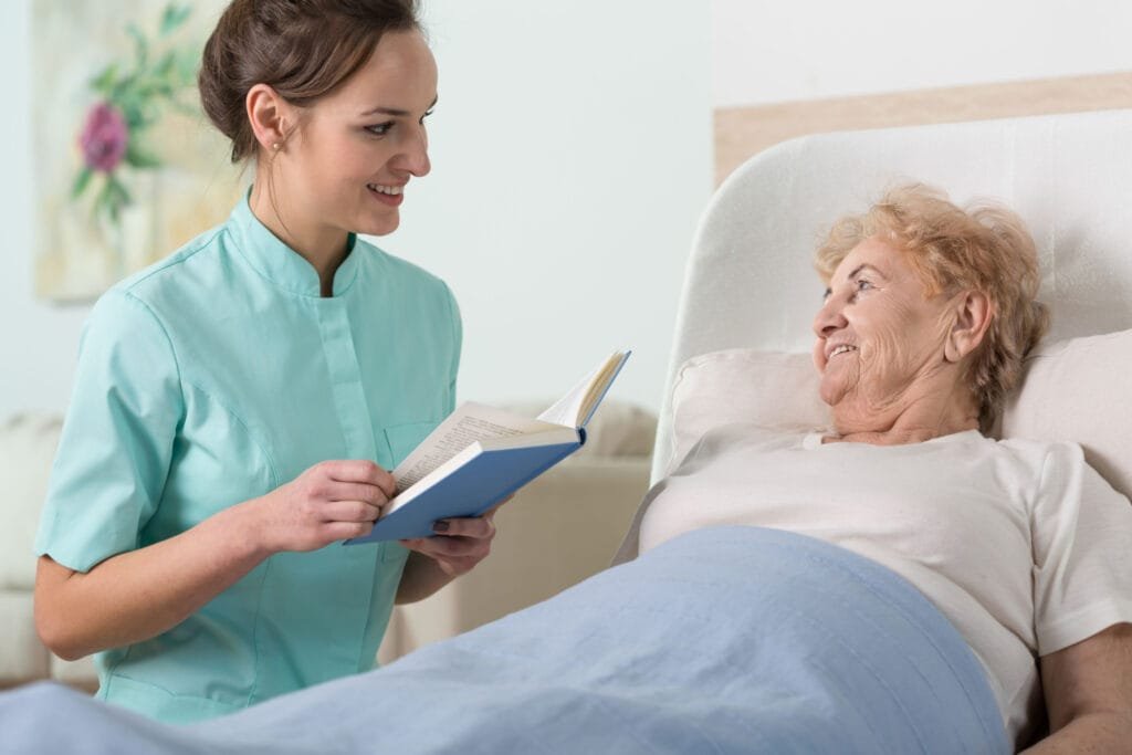 Young beautiful nurse reading to sick older woman in bed